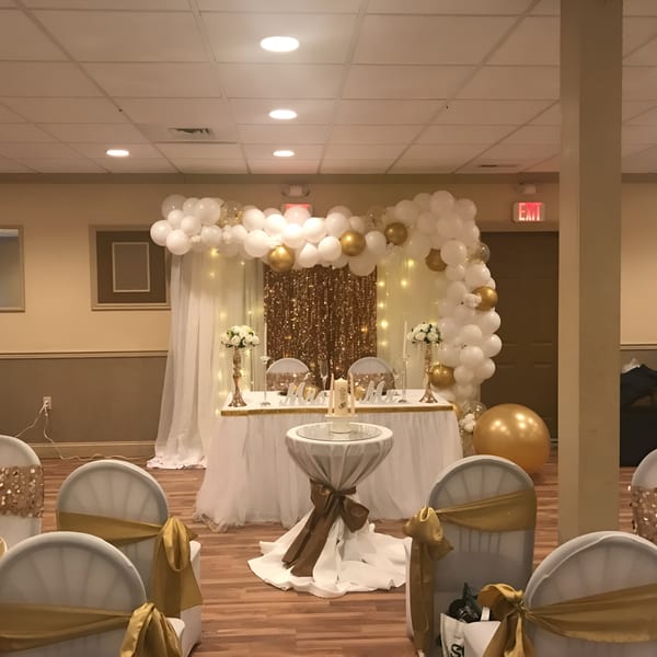 Elegant banquet hall decorated for a wedding with white and gold balloons, draped tables, and chairs.