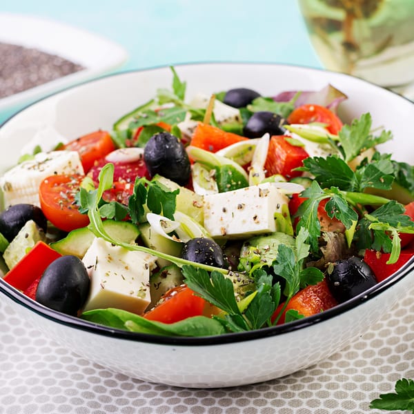 Close-up of a vibrant Italian salad with feta, olives, tomatoes, cucumbers, and herbs in a white bowl.