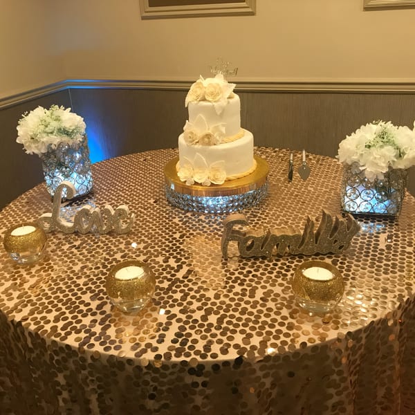 A two-tier white cake with peach roses on a gold sequin tablecloth, flanked by floral arrangements.