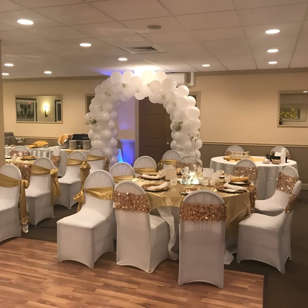 Banquet hall set for an event with white chairs, gold table linens, and a white balloon arch.
