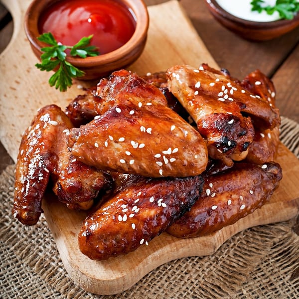 Glazed chicken wings with sesame seeds, ketchup, and a white dipping sauce on a wooden board.
